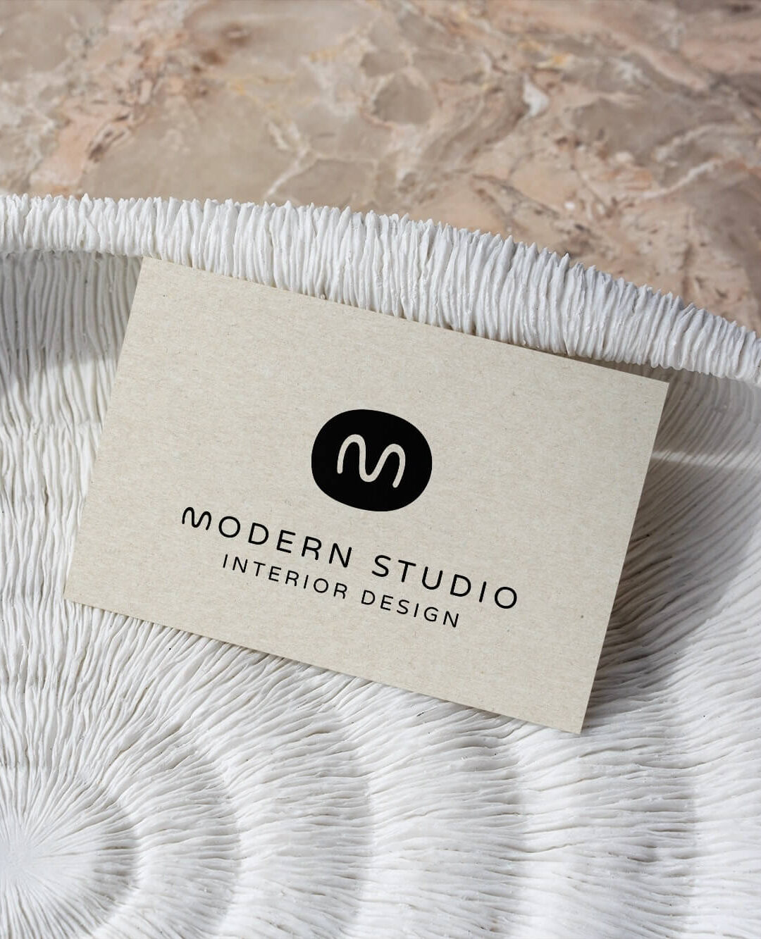 A business card for "Modern Studio Interior Design" rests on a textured white bowl with a spiral design. The card features a minimalist logo with a black circle displaying a white "M" and the text in black below. The background is a marble surface.