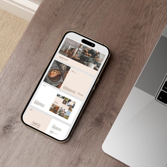 A smartphone displaying a brand guide for 'salt & Chocolate' with images of cakes and pastries lies on a wooden desk next to a partially visible laptop. The desk also has a light-coloured carpet at the edge.