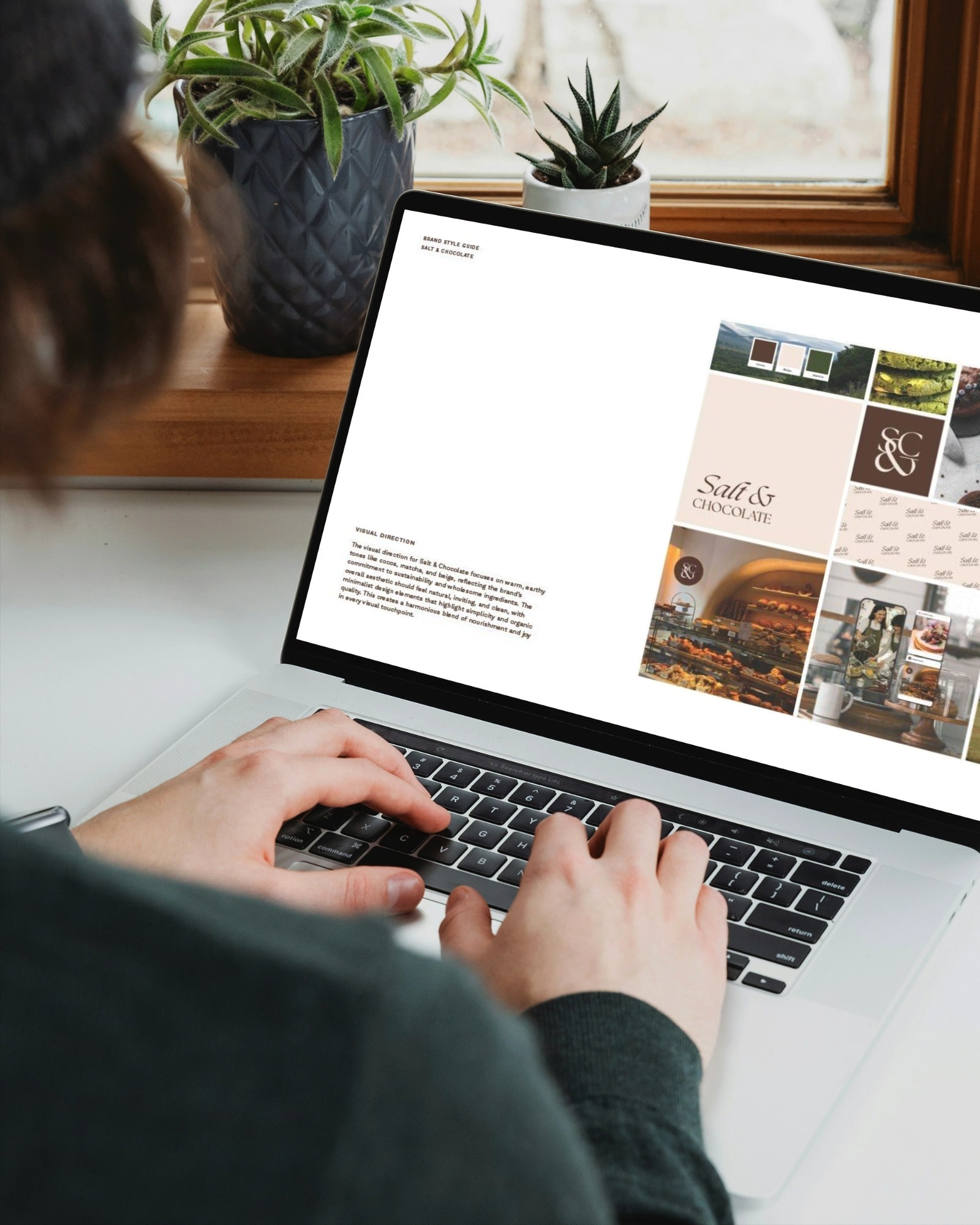 A person types on a laptop displaying a design or presentation document featuring images and text. The screen shows a grid layout with various graphics and a section of text, adhering to brand guidelines. There are potted plants and a window in the background, and the person wears a dark top.