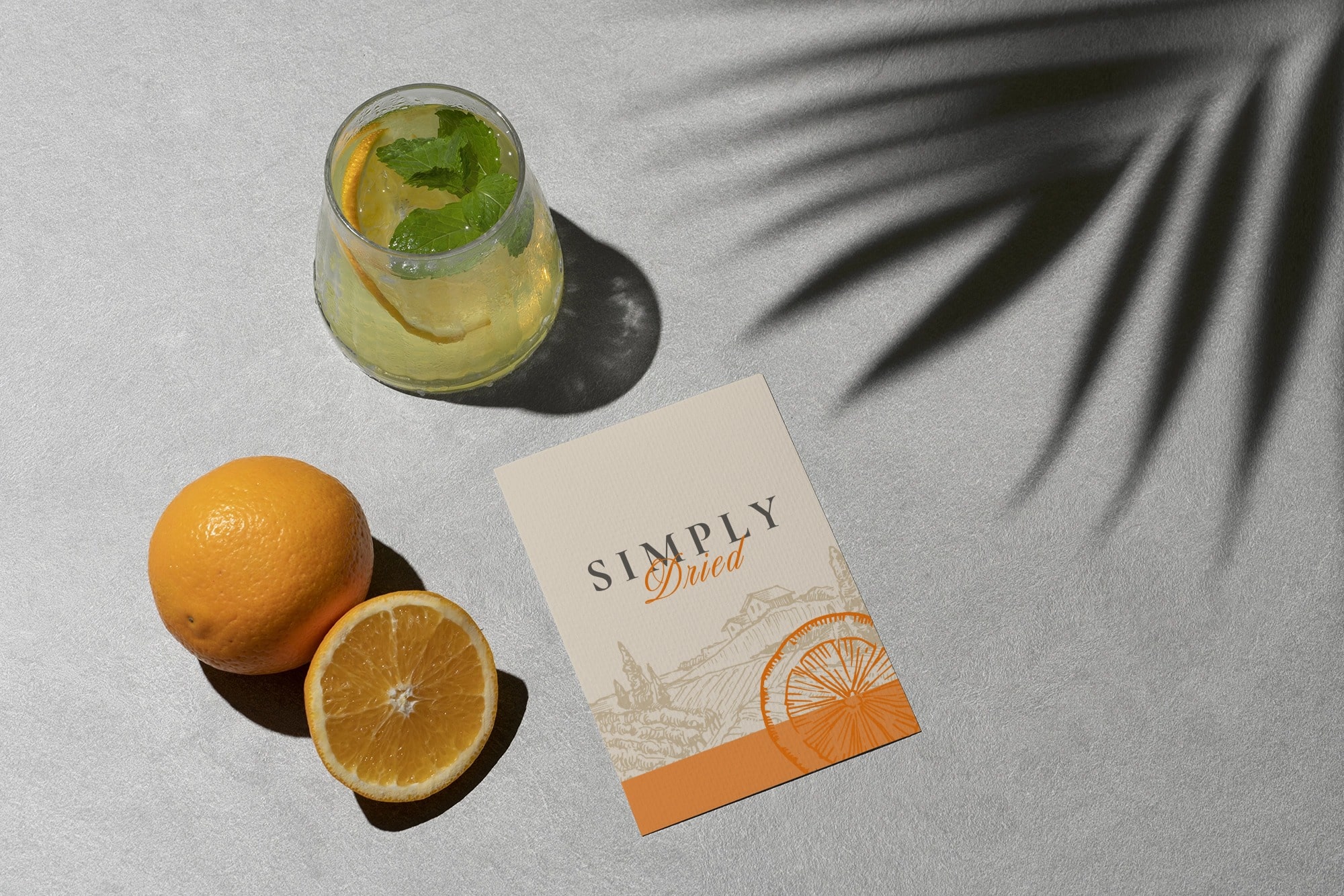 A refreshing drink with mint leaves in a glass is placed next to halved and whole oranges on a gray surface. A card with "Simply Dried" written on it, showcasing elegant branding, lies nearby. The shadow of a palm leaf casts on the surface, creating a tropical ambiance perfect for print collateral.